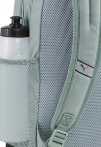 Hele roheline seljakott, millel on võrkturv ja valge veepudel. Sujuv kangas ja must Puma logo rihmal.