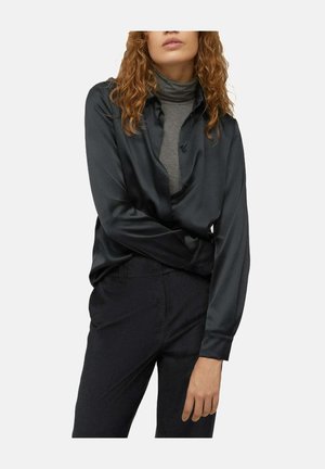 Donna che indossa una camicia scura di seta a maniche lunghe sopra un dolcevita grigio e pantaloni neri, in piedi con le braccia incrociate e capelli ricci visibili.