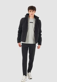 Giacca softshell nera con cappuccio, T-shirt grigia con una striscia di logo frontale e pantaloni neri. Il modello indossa scarpe da ginnastica chiare.