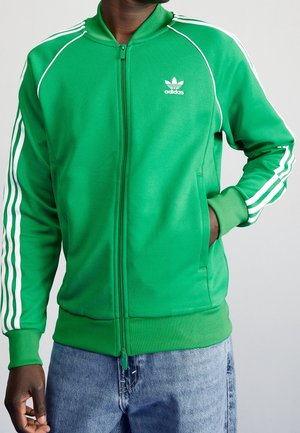 Homme portant une veste de survêtement zippée verte Adidas avec des bandes blanches sur les manches et un logo sur la poitrine, associée à un jean bleu, main dans la poche.