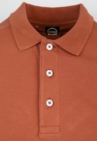 Polo a colletto color ruggine con tessuto testurizzato, tre bottoni bianchi e un sottile motivo intrecciato sul colletto.