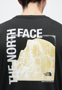 Μαύρο βαμβακερό μπλουζάκι με μεγάλο λευκό και κίτρινο γραφικό ενός βουνού και το κείμενο και το λογότυπο "THE NORTH FACE" στην πλάτη.