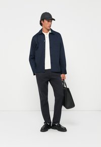 Man wearing black cap, navy jacket over cream polo, black pants, and black shoes, holding a black leather tote bag against a plain white background.