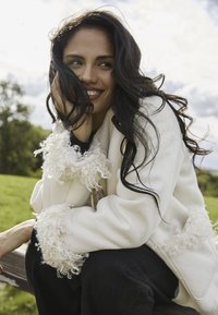 Biała kurtka z puszystymi mankietami, czarna bluzka i ciemne dżinsy. Długie ciemne włosy opadają na ramiona, siedzi na drewniej ławce.