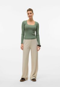 Haut long à manches ribbé vert avec un décolleté carré, associé à un pantalon large beige et des chaussures marron foncé, présentant une texture lisse.