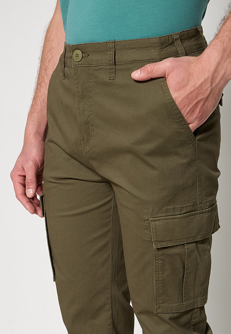 Mężczyzna noszący oliwkowe spodnie typu cargo z ręką w prawej kieszeni oraz turkusową koszulę wsuniętą do spodni.