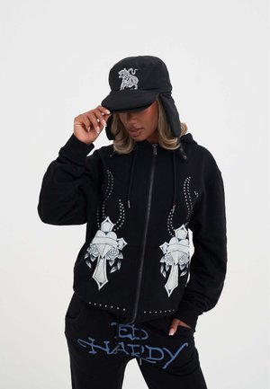 Felpa nera con zip, decorata con motivi floreali e borchie sul davanti, abbinata a pantaloni neri con lettering blu. Berretto isolato nero indossato.