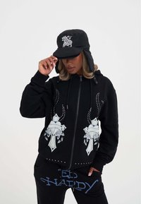 Felpa nera con zip, decorata con motivi floreali e borchie sul davanti, abbinata a pantaloni neri con lettering blu. Berretto isolato nero indossato.