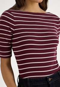 Mulher vestindo uma blusa ajustada com riscas horizontais em bordô e branco, com mangas até ao cotovelo, enfiada em jeans escuros de ganga.