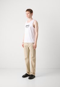 Witte katoenen tanktop met een zwart grafisch logo, gecombineerd met lichtbeige broek en zwarte schoenen, staand op een lichte achtergrond.