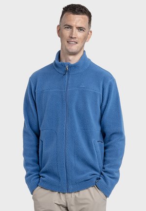 Mann trägt eine blaue Fleecejacke mit durchgehendem Reißverschluss, die Hände in den Taschen, lächelt sanft vor einem schlichten hellen Hintergrund.