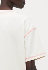 T-shirt di colore crema con una vestibilità rilassata, caratterizzata da cuciture rosa lungo il colletto e le maniche. Tessuto morbido con una texture liscia.