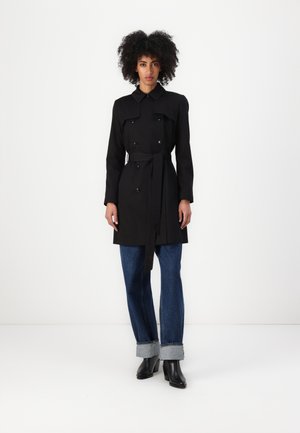 Zwarte trenchcoat met een dubbel knoopdesign, voorste knopen en een strikceintuur. Draagt over een blauwe denim jeans met opgerolde mouwen.