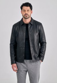 Mann mit Bart, der eine schwarze Lederjacke über einem schwarzen Hemd und grauen Hosen trägt, steht mit einer Hand in der Tasche vor einem einfarbigen Hintergrund.