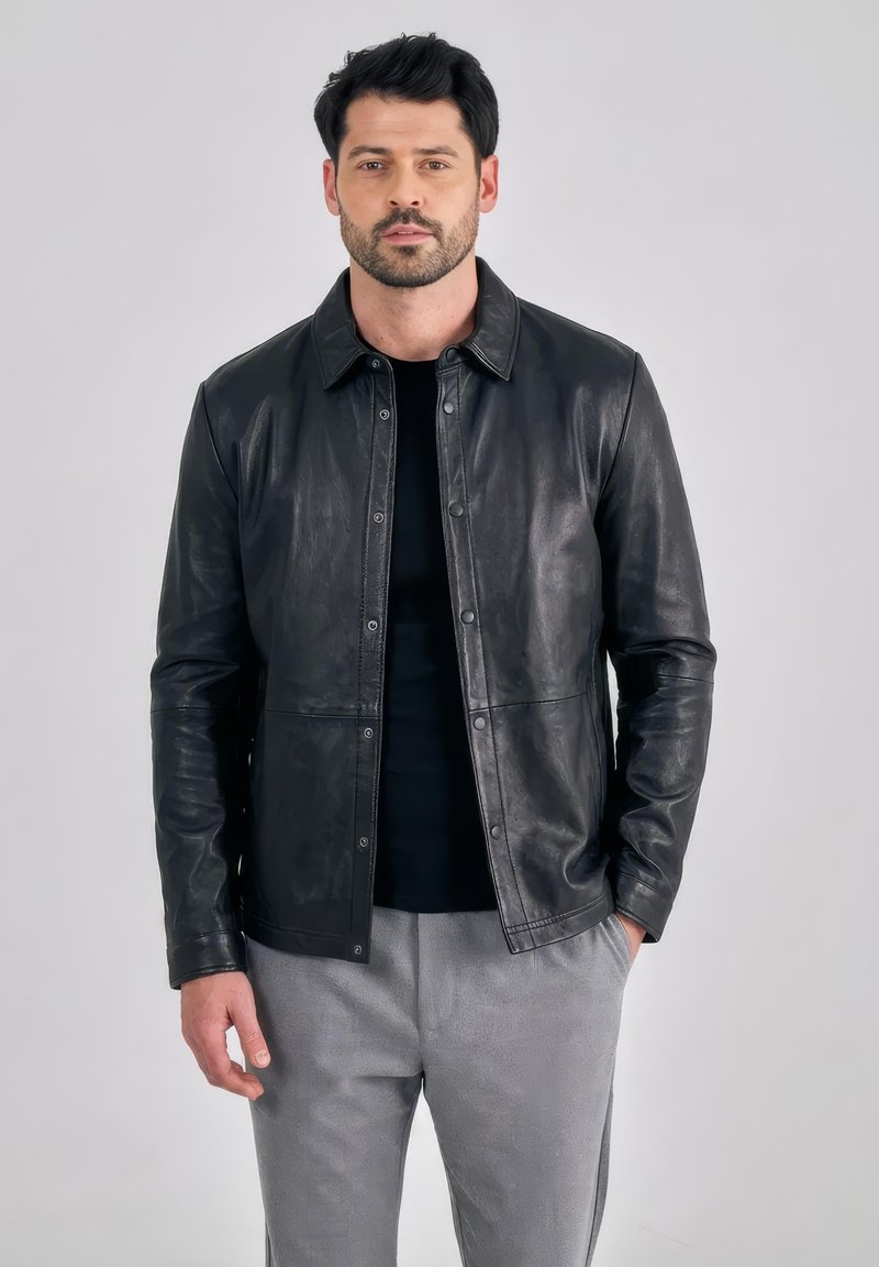 Mann mit Bart, der eine schwarze Lederjacke über einem schwarzen Hemd und grauen Hosen trägt, steht mit einer Hand in der Tasche vor einem einfarbigen Hintergrund.