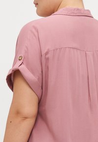 Chemise rose clair à manches courtes avec col, présentant des manches roulées et un détail plissé dans le dos. Tissu lisse avec un léger éclat.