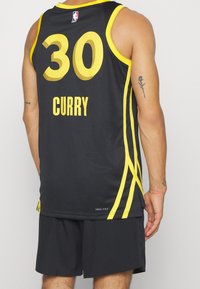 Henkilö, joka käyttää mustaa ja keltaista koripallopaitaa, jossa on numero 30 ja nimi Curry, yhdistettynä mustiin shortseihin, ja näyttää käsivarsi-tatuoinnit.