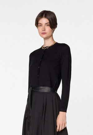 Frau trägt eine schwarze, geknöpfte Strickjacke, einen schwarzen plissierten Rock mit Ledergürtel und eine grobe goldene Gliederkette, steht vor weißem Hintergrund.