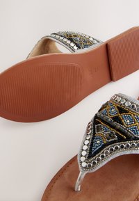 Tongs ornées de perles avec une tige à motif en bleu, or et argent. Semelle marron texturée à design plat et brides transparentes.