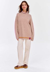 Beige Pullover mit pinken Akzenten, hohem Kragen und Seitenschlitzdetails. Kombiniert mit cremefarbenen Hosen und braunen Sneakers mit pinken Streifen.