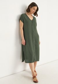 Grünes, knielanges Kleid mit kurzen Ärmeln, V-Ausschnitt und Spitzenverzierung vorne. Leichter Stoff mit Seitenschlitzen, kombiniert mit braunen Sandalen.