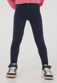 Granatowe legginsy wykonane z elastycznego materiału, dopasowane w całej długości nóg, zestawione z czarnymi sneakersami za kostkę z białymi akcentami i różowymi podeszwami.