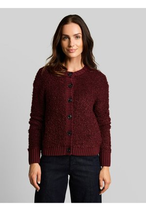 Vrouw met lang bruin haar die een bordeauxrode textuurige getailleerde vest met knopen en een donkere broek draagt, staand tegen een grijze achtergrond.