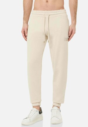 Man wearing beige jogger pants with drawstring waist and white sneakers with black accents, standing against white background.