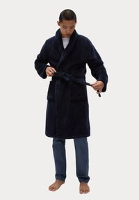 Granatowy, pluszowy szlafrok z szalem, z paskiem w talii oraz dwiema przednimi kieszeniami. Model ma na sobie jasne spodnie, stoi na boso.