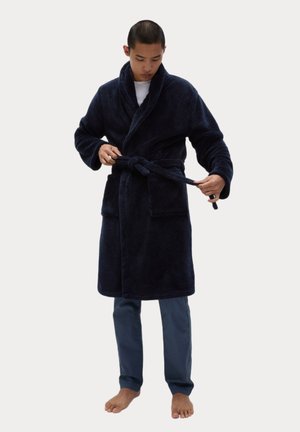 Navyblauwe, pluche badjas met een sjaalkraag, een ceintuur in de taille en twee voorkeurszakken. Model draagt lichte broek, staat blootvoets.