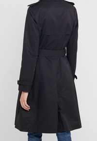 Svart trench coat med bälte i midjan, axelknappar och knäppta ärmar. Har en slät, texturerad tyg och är medellång i designen.