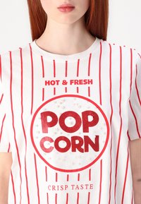 Valkoinen t-paita, jossa on punaiset pystysuorat raidat. Siinä on pyöreä "POP CORN" -kuvio kimaltavalla pinnalla, jota korostavat pienet valkoiset helmet ja teksti "HOT & FRESH."