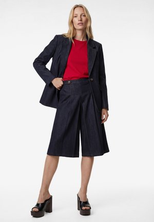Vrouw draagt een rood topje, donkerblauwe knie-lange wijdere broek, bijpassende blazer en zwarte platform open-toe hakken, staand tegen een witte achtergrond.