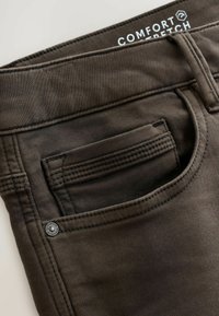 Detailný záber na tmavohnedú kapsu na nohaviciach s viditeľným šitím, kovovým nitom a čiastočnou bielou značkou "Comfort Stretch".