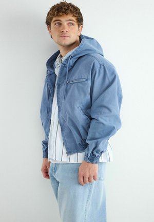 Mladý muž s modrou kapucňou na bielej pruhovanej košeli a svetlo modrými džínsami, ktorý sa pozerá doľava na jednofarebnom pozadí.