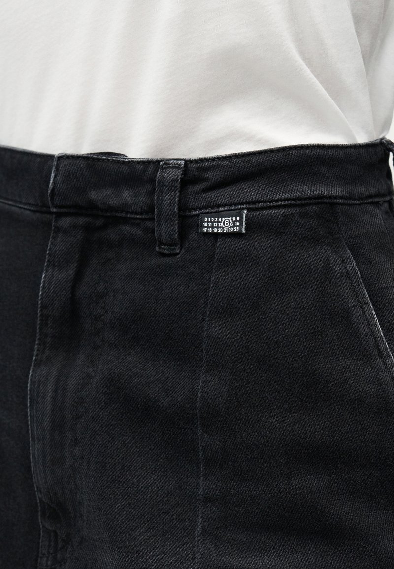 Pantaloni in denim neri a vita alta con un'etichetta bianca con numeri, indossati con una camicia bianca infilata.