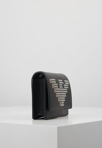 Černá kožená peněženka s stříbrným a zlatým hřebíčkovaným symbolem eura na přední straně, stojící vzpřímeně na bílé ploše proti jednoduchému pozadí.