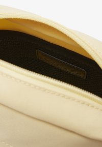 Gelber Reißverschlussbeutel mit strukturierten Oberflächen; schwarzes Netzfutter sichtbar und ein kleines "LACOSTE"-Etikett innen. Glatter Reißverschluss und Nähte.