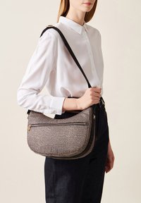 Borsa a tracolla a pois bianco e nero, di forma rotonda, con tasca con zip e tracolla regolabile, indossata su una camicia bianca.