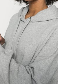 Primer plano de una persona que lleva una sudadera ligera gris con gruesos cordones y un pequeño logo bordado en el pecho, con los brazos cruzados.