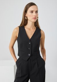 Crni prsluk s dubokim V-izrezom, zatvaranjem na četiri dugmeta i bočnim džepovima. Prilagođeni kroj s glatkom tkaninom i bez vidljivih uzoraka.