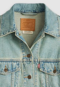 Lichtblauwe spijkerjacket met een klassieke kraag, bronzen knopen en een leren label met merkdetails. Gelaagde stof met contrasterende stiksels.