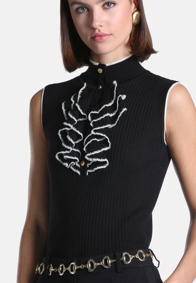 Femme portant un haut sans manches noir côtelé avec détail de volants blancs et boutons dorés, associé à une ceinture chaîne dorée et de grandes boucles d'oreilles dorées.