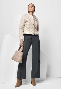 Beige gewatteerde jas met zwarte knopen, gecombineerd met grijze wijdvallende broek en bruine hiel enkellaarsjes. Kleine beige handtas inbegrepen.