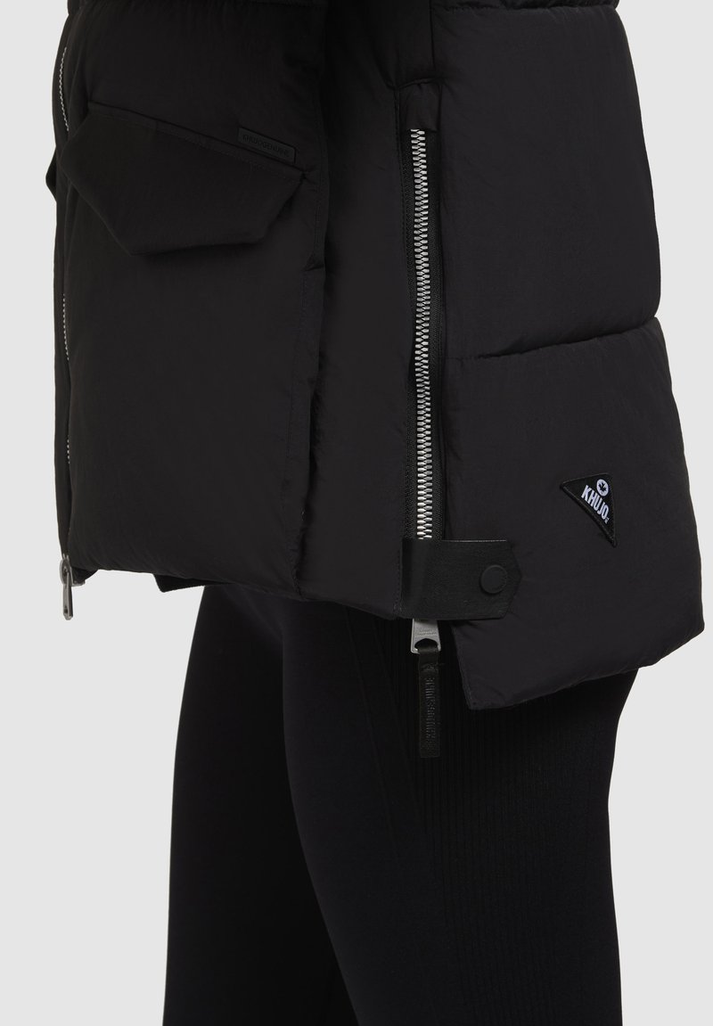 Schwarze Pufferjacke mit glänzender Textur, mit Reißverschlusstaschen und Logopatch. Hat ein tailliertes Design und verstellbare Hardware-Details.