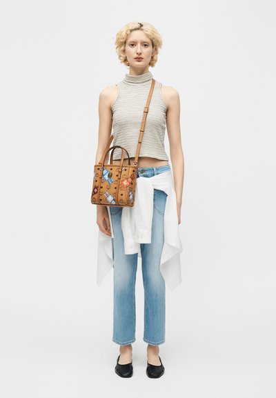 Νεαρό άτομο με κοντά σγουρά ξανθά μαλλιά, φορώντας αμάνικη ριγέ μπλούζα, ανοιχτόχρωμα μπλε τζιν, μαύρες φλατ, λευκό πουκάμισο δεμένο στη μέση, κρατώντας καφέ τσάντα με σχέδια.