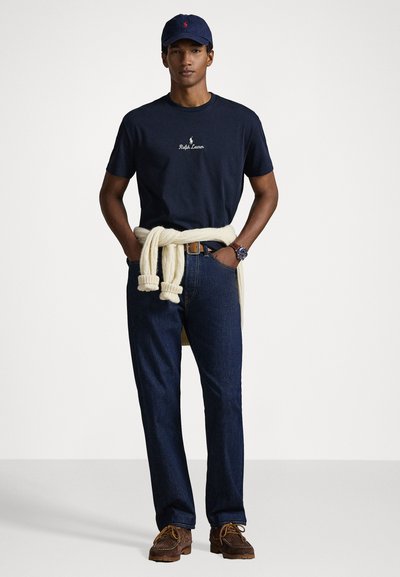 T-shirt azul marinho com o logótipo "Polo Ralph Lauren", suéter creme amarrado à cintura, jeans escuros, mocassins castanhos e boné azul marinho. Estilo de roupa casual.