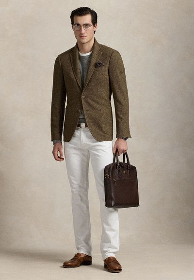 Hombre con blazer marrón, suéter gris, pantalones blancos y zapatos marrones, sosteniendo un maletín de cuero marrón oscuro en la mano derecha.