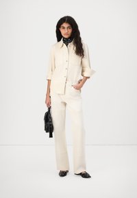 Beige denim shirt and matching high-waisted pants with side pockets. Black handbag and loafers, featuring round cutouts. Neutral background.