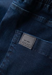 Dunkelblauer Denimstoff mit feiner Textur, versehen mit einem quadratischen Lederpatch, das mit "ombre" und "EST. 1985" beschriftet ist und in die Gesäßtasche eingenäht ist.